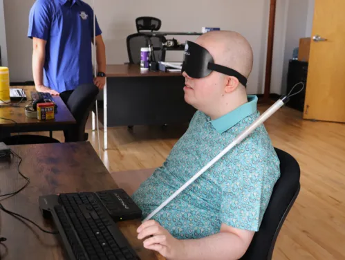 A man in sleep shades sits at a desk using a braille display and keyboard.