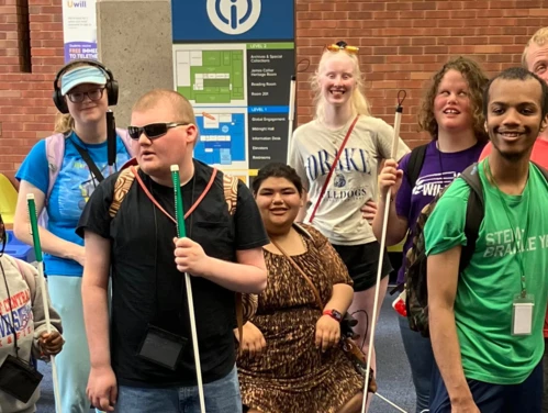 A group of students holding white canes stands together in front of a brick-walled indoor space.