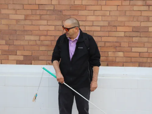 A man stands outdoors in front of a brick wall, holding a long white cane.