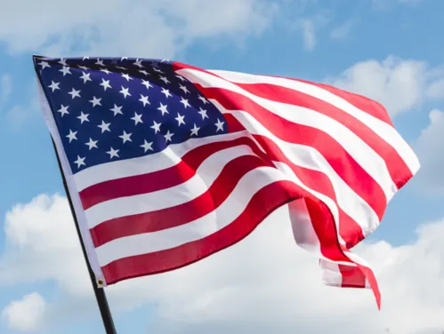 A close shot of an American flag blowing in the wind. Behind it is a blue sky with clouds.