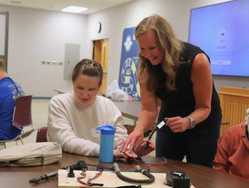 A woman is standing by a female student helping her plug wires into a board