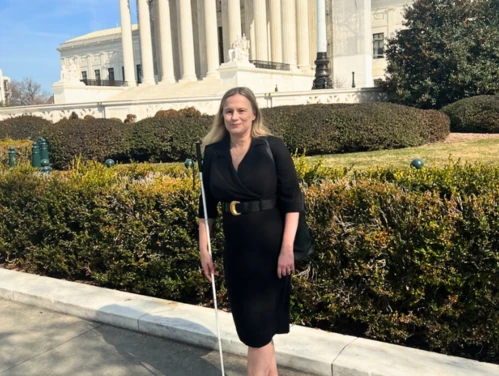 A woman stands outdoors in front of a large white building with tall columns. She is wearing a black dress with a belt, carries a black bag over her shoulder, and holds a long white cane.