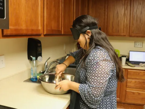 A woman wearing sleep shades is standing in a kitchen stirring something in a large metal bowl