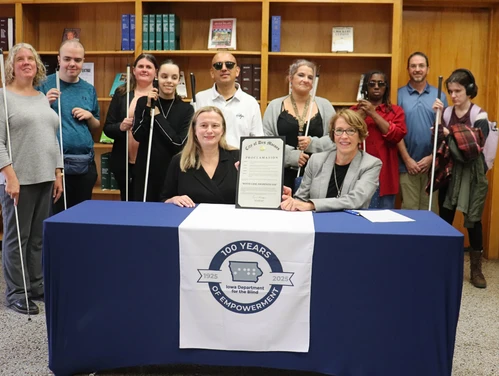 A group of people, some holding white canes, stand behind a table draped with an Iowa Department for the Blind centennial banner as two women seated in front hold a White Cane Awareness Day proclamation.