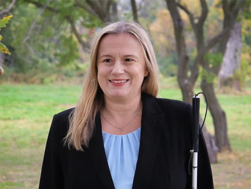 Woman with long blonde hair smiles in a park, wearing a black blazer and holding a white cane.