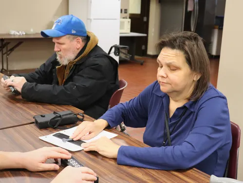 Two adults sit at a table during a braille lesson. A woman uses a slate and stylus to take notes.