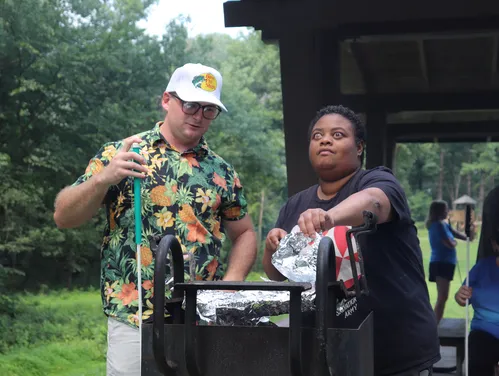 A man and woman stand at a grill outside, as the woman uses tongs to remove aluminum foil from the grill