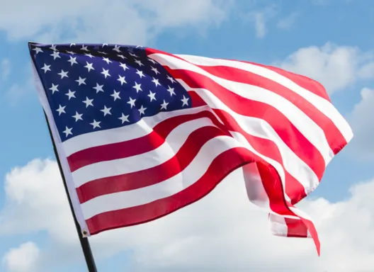 A close shot of an American flag blowing in the wind. Behind it is a blue sky with clouds.