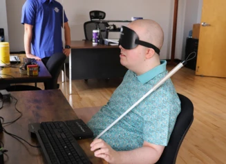 A man in sleep shades sits at a desk using a braille display and keyboard.