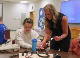 A woman is standing by a female student helping her plug wires into a board
