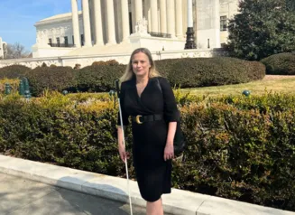 A woman stands outdoors in front of a large white building with tall columns. She is wearing a black dress with a belt, carries a black bag over her shoulder, and holds a long white cane.