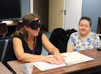 A woman in sleep shades sits at a table reading print braille while another woman sits beside her smiling