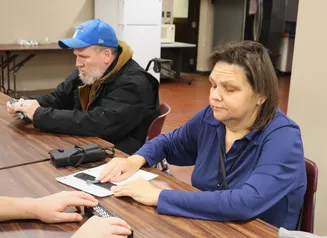 Two adults sit at a table during a braille lesson. A woman uses a slate and stylus to take notes.