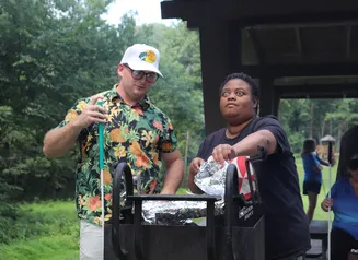A man and woman stand at a grill outside, as the woman uses tongs to remove aluminum foil from the grill