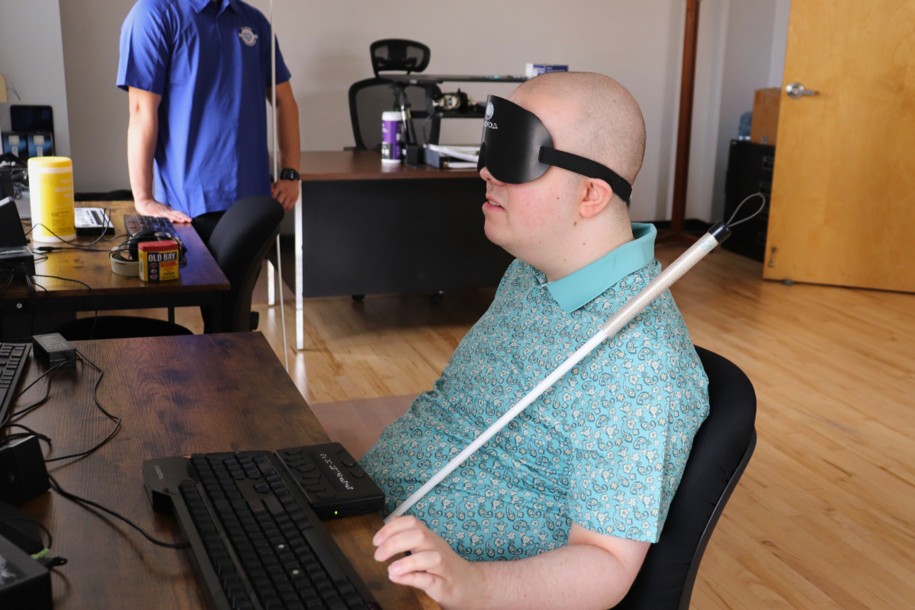 A man in sleep shades sits at a desk using a braille display and keyboard.