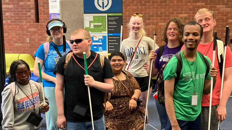A group of students holding white canes stands together in front of a brick-walled indoor space.