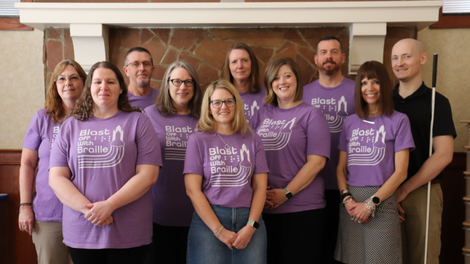 A group of ten people stands together indoors in front of a stone fireplace. Most of them are wearing matching purple t-shirts that read "Blast Off with Braille" 