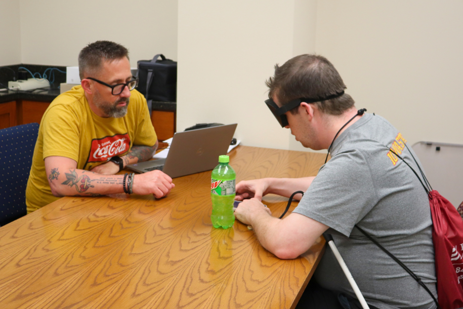 Two people seated at a table. One is using a laptop, while the other is using his phone.